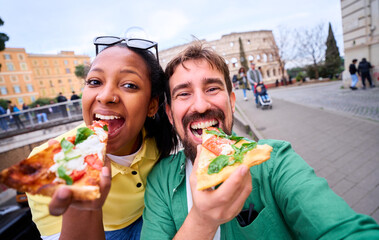 Happy mixed race couple taking a fun selfie while eating pizza in front of colosseum in Rome. Image with copy space.