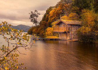 Charming stone boathouse on a still lake surrounded by colourful autumn trees in the Lake District, England.