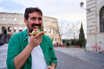 Fototapeta premium Cheerful man eating a slice of pizza with the iconic Colosseum in the background, immersed in a delightful culinary experience in Rome. Copy space.