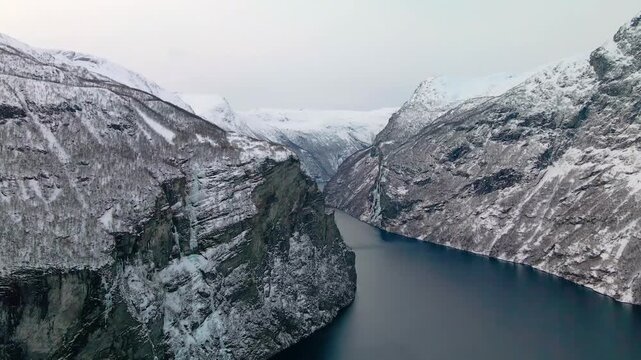 This drone footage shows Geirangerfjord in winter, a UNESCO World Heritage site in Norway. The serene and dramatic landscape of snow capped mountains, steep cliffs, and the deep blue waters of the fjo