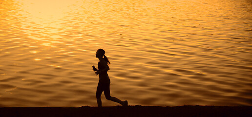 black silhouette of one Alone young sporty girl athlete running at sunset sea river along horizon line hill beach Female lady hold in hand mobile cell phone and listen powerful energy music Sexy body