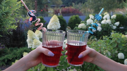 aperitivo all'aperto in giardino pieno di fiori, le mani femminili che tengono bicchieri pieni con una bevanda di colore rosso