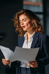 Young professional woman holding sheets of paper, rehearsing a speech in a modern corporate office, preparing for a presentation, job interview, or public speaking event. 