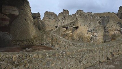 Herculaneum, Italy - 9 January 2025. Stone ruins of Roman-era buildings with mosaic-patterned walls and remains of entrances and passageways under cloudy skies.