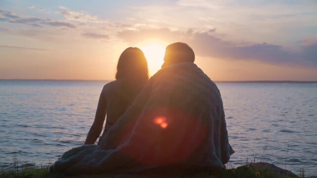 A young couple sits at sunset in warm backlight as the man gently covers the woman with a blanket, creating a romantic cozy mood.