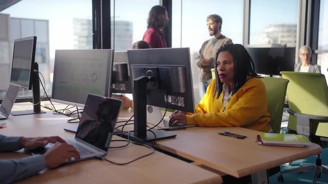 Focused african millennial woman in yellow sweater using laptop in a bright office with colleagues, teamwork atmosphere, modern business workspace, city view - Powered by Adobe