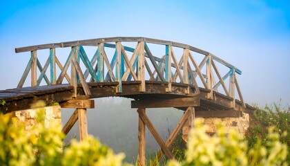 A weathered wooden bridge arches across a misty landscape, its aged blue paint contrasting against the light wood.
