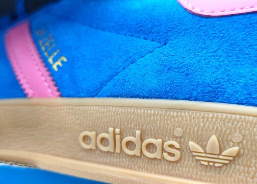 Closeup view of adidas Gazelle sneakers in blue suede with pink leather stripes and heel tab, featuring a classic gum sole and gold branding for a bold retro style.