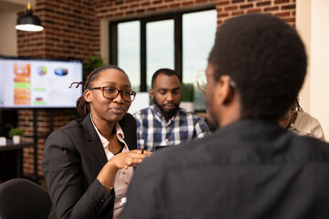 Female manager talks through marketing strategies with black male employee, asking questions and analyzing financial report. Businesswoman discussing project updates with colleague in modern office.