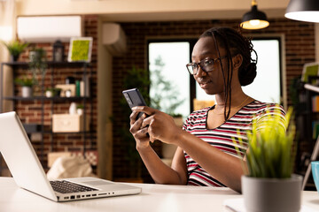 African american female entrepreneur looking at phone screen in astonishment after receiving surprising text. Young black woman seated at desk with laptop, reacting to unexpected news on mobile device