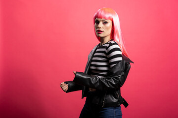 Gorgeous woman with striped crop top stands facing camera under soft lighting with vibrant hairstyle and radiant skin. Young model in black jacket conveys calm elegance, posing against pink background