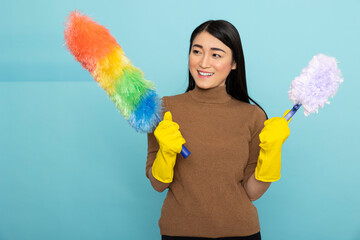 Happy asian woman wearing yellow rubber gloves holds rainbow and white duster, smiling confidently while posing for portrait against blue background. Housekeeping and hygiene concept.