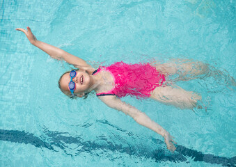 Young girl wearing goggles and a pink swimsuit enjoying backstroke in bright blue pool water, radiating joy and carefree summer fun