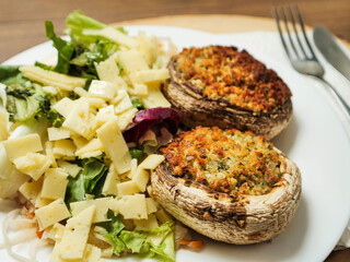 A plate of food with a fork and knife on a wooden table. The plate has a fresh salad with cheese and two fancy posh stuffed mushroom patties. Simple tasty meal.