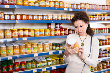 Female shopper selects glass jar of sauerkraut at a grocery store. Woman shopper reading expiration date on product label