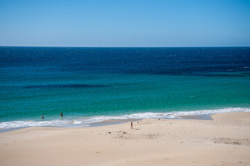 Wide sandy beach with turquoise ocean and people swimming under blue sky
