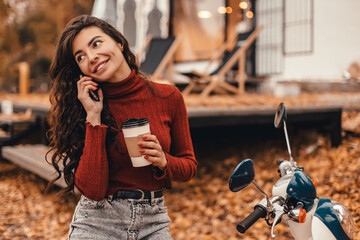 Beautiful woman in cozy outfit using smartphone or phone and drink coffee while sitting on chair near cafe in autumn park. Freelancer works remotely outside home.