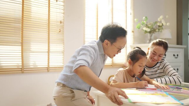 Happy Asian family parents teaching little child girl do homework in living room. Father, mother and daughter enjoy spending time together at home. Family relationship, homeschool student education.