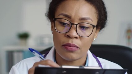 A focused doctor with glasses writing notes on a clipboard - Powered by Adobe