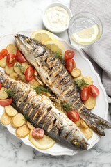 Tasty roasted seabass fish served with potatoes, tomatoes, sauce and lemon on white marble table, flat lay. Homemade seafood dish
