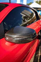 Car side mirror with a carbon fiber cover and integrated turn signal repeater, on a vehicle with red paint.