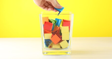 Woman picking light blue paper piece from glass jar at wooden table against yellow background, closeup