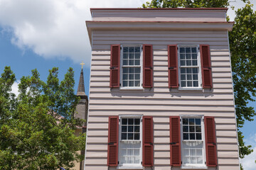 Charleston, South Carolina, USA - July 2, 2025: A pastel pink historic house with bold red shutters and sash windows, with a church steeple visible behind.