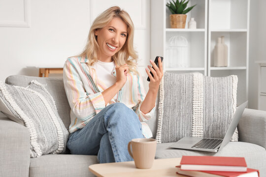 Happy mature woman using mobile phone on sofa at home - Powered by Adobe