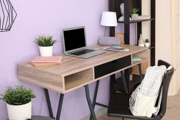 Modern school desk with stationery and laptop in room near purple wall