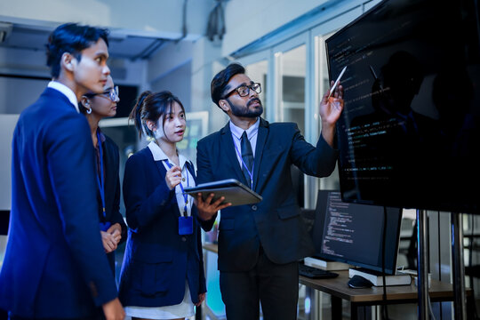 A senior AI architect leads a training session for a diverse team of developers. He explains a complex machine learning algorithm, pointing to code on a large vertical monitor.