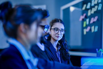 A female AI team lead guides a developer in an agile scrum session. She points at the screen, collaborating on a complex machine learning task with a kanban board in the background.