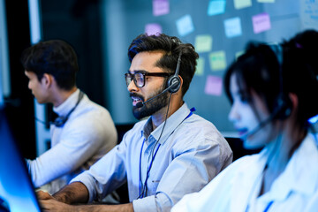 Professional call center agents with headsets providing friendly customer support. A dedicated male operator and his team work in modern all day technical assistance center.
