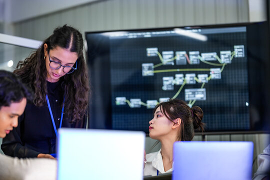 A diverse group of professionals in a corporate workshop on AI development. A female instructor supervises as the team learns to code advanced machine learning models on their laptops.