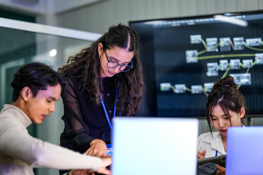 A diverse group of professionals in a corporate workshop on AI development. A female instructor supervises as the team learns to code advanced machine learning models on their laptops.