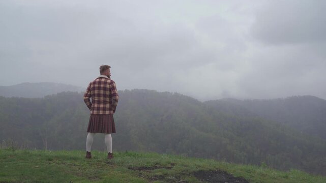 An adult redhead bearded man in Scottish national dress plaid jacket and kilt stands on a hill and looks into the distance. Masculinity, pride and human dignity concept.v