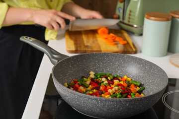 Woman putting cut carrot into frying pan on stove in kitchen, closeup