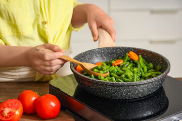 Woman with spoon frying vegetables on mini stove in kitchen, closeup