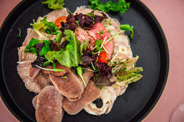 A variety of meat snacks, boiled beef tongue, roast beef and meat roll on a black plate with lettuce leaves and microgreens. Restaurant dish on a light pink tablecloth