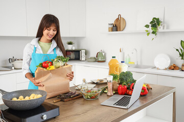 Young woman with paper bag of food and laptop in kitchen