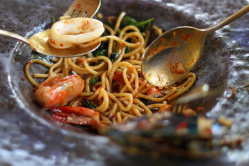 Close Up view of seafood basil spaghetti on a plate, featuring a spicy Thai flavor. It has a spoon and fork and squid as ingredients.