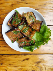 A top-down view of a delicious-looking plate of fried eggplant slices topped with a garlic sauce and herbs. The dish is served with fresh lettuce on a white plate, placed on a rustic wooden table.