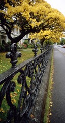 Obraz premium Ornate black wrought iron fence borders a lush green lawn, framed by a vibrant yellow autumn tree canopy, in front of charming light-colored houses.