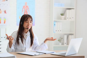 Female Asian doctor with headset and laptop giving online consultation in clinic