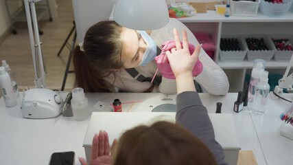 Top view of nail technician in mask applying brush to client nail under desk lamp while manicure tools, brushes, and bottles are arranged on table during nail care session in beauty salon