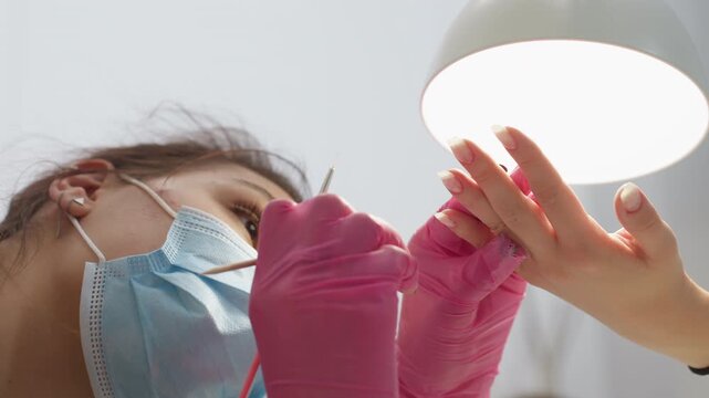 Nail technician in face mask and pink gloves carefully applies hardener to client nails under bright lamp, focusing on precision and detailing each fingernail while client hand is gently held