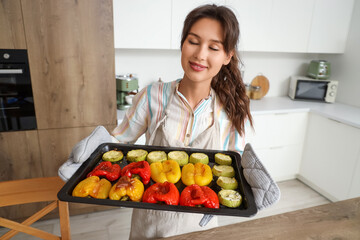 Young woman holding baking tray with vegetables in kitchen