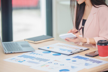 Smiling young Asian businesswoman is thinking and analyzing work plan in office hard using laptop, smartphone, calculator and documents. Chart sheet on table. Business and finance concept.