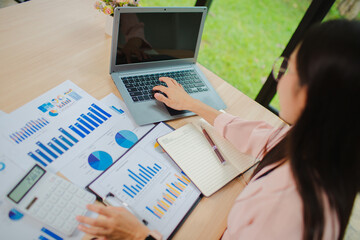 Smiling young Asian businesswoman is thinking and analyzing work plan in office hard using laptop, smartphone, calculator and documents. Chart sheet on table. Business and finance concept.