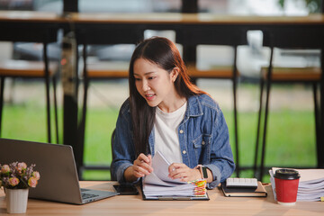 Smiling young Asian woman clearing paperwork in office, using laptop and many documents beside her. Accounting and finance business concept.