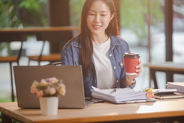 Smiling young Asian woman clearing paperwork in office, using laptop and many documents beside her. Accounting and finance business concept.